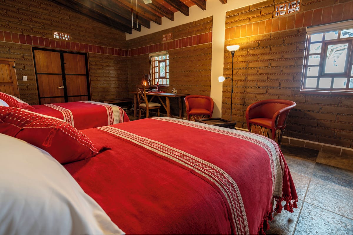 A room with a wooden wall, a bed with red bedding, a table with chairs, and a window with a view.