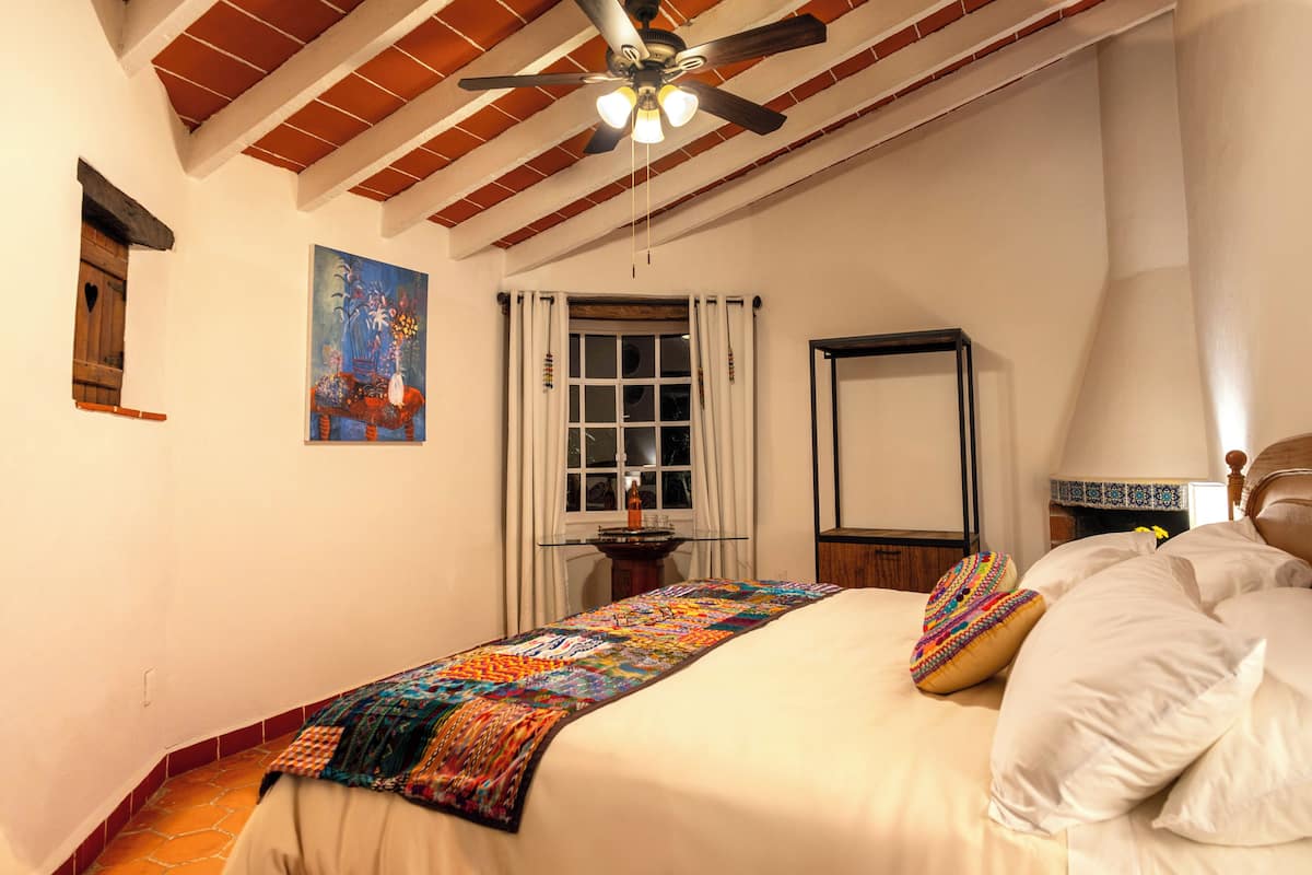 A bedroom with a bed, a ceiling fan, a window with curtains, a painting on the wall, and a small table with a lamp.
