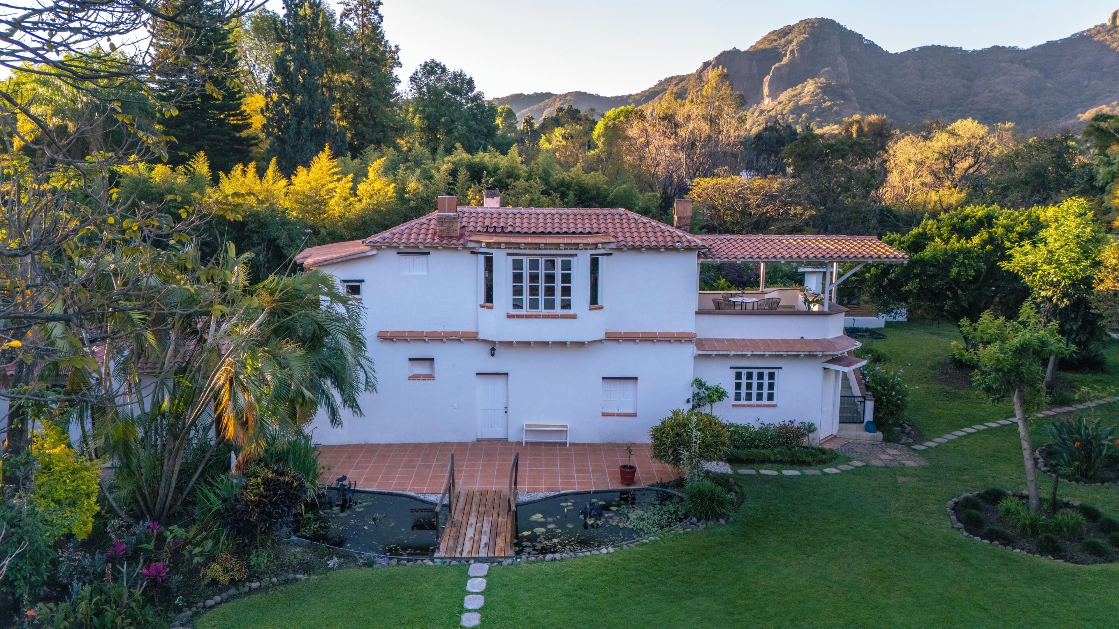 A two-story house with a tiled roof, surrounded by lush greenery and a small pond.