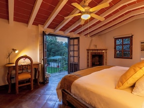 A bedroom with a bed, a desk with a chair, a ceiling fan, a fireplace, and a view of the garden. - LUZ KAUANI (Tepoztlan)