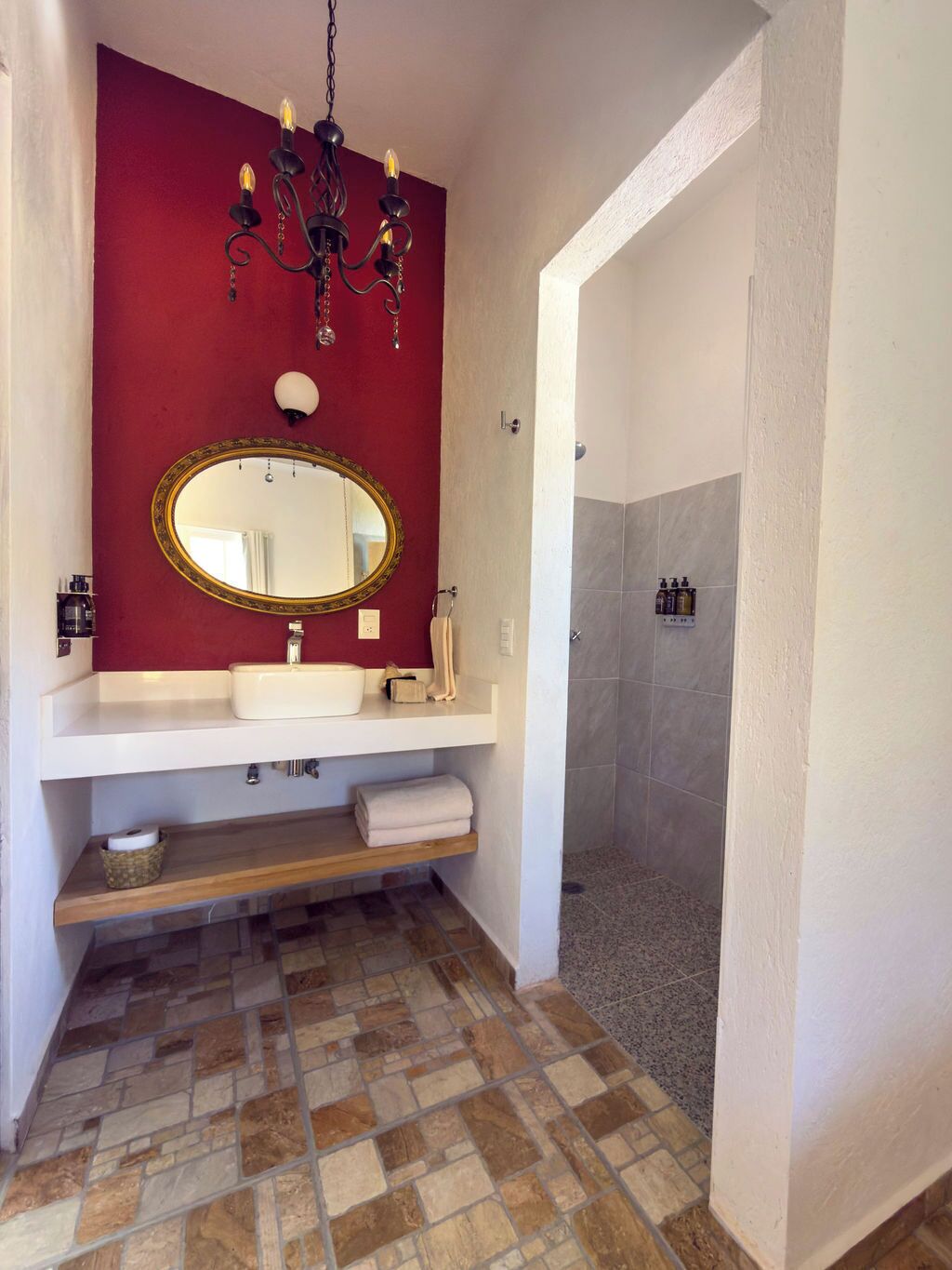 Een badkamer met een rode wand, een wit wastafelmeubel, een spiegel met gouden lijst, een kroonluchter en een douchegedeelte.