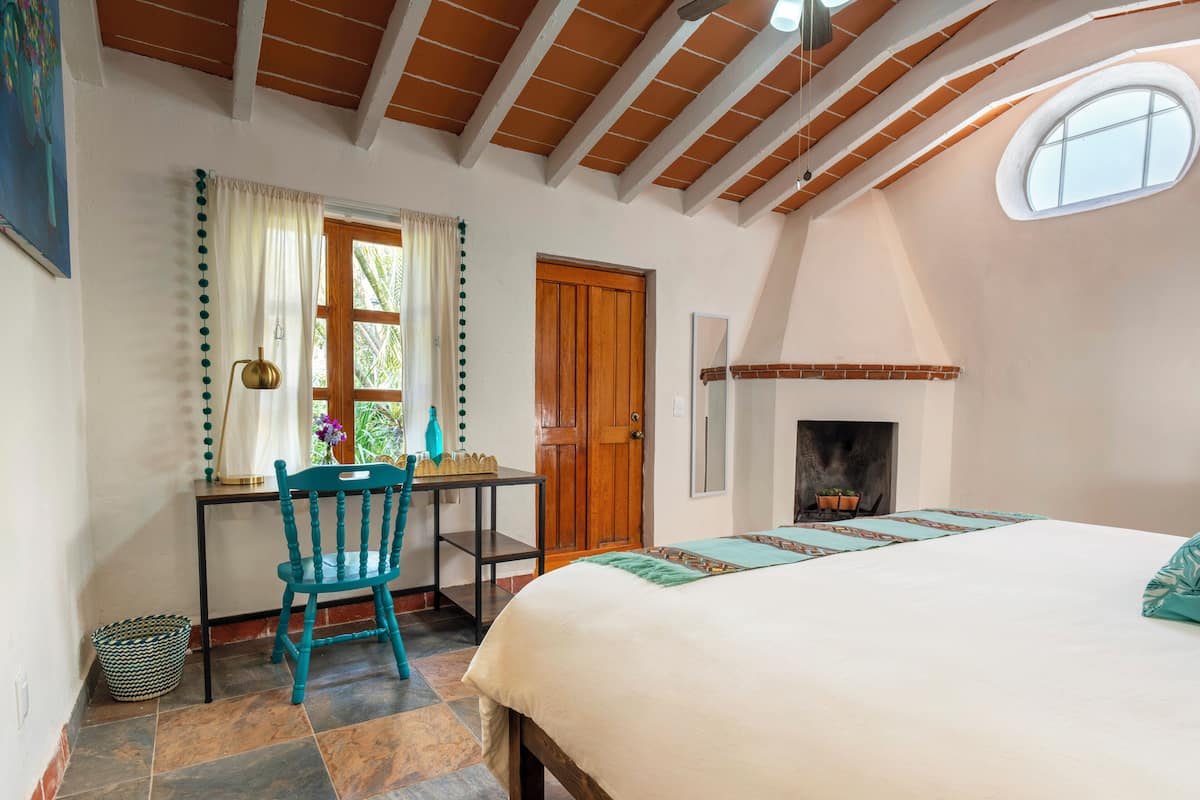 A bedroom with a bed, a desk, a chair, a fireplace, and a window.