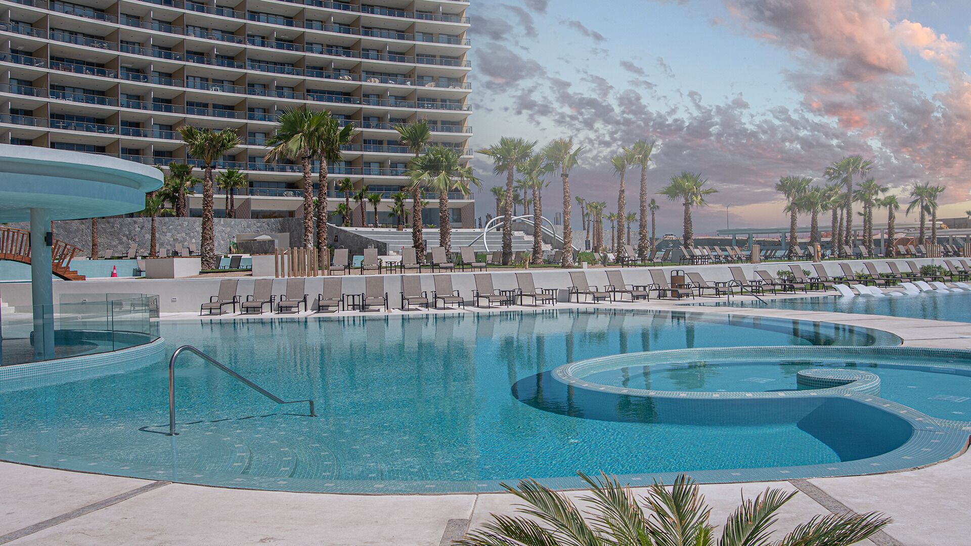 Piscina de resort con sillas de descanso y palmeras, rodeada por un edificio de hotel moderno.