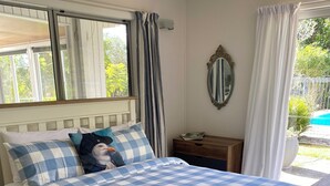 A bedroom with a checkered bedspread, a wooden headboard, a nightstand, a mirror, and a view of a swimming pool. - Mission Retreat - spacious and peaceful (Kerikeri)