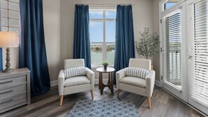 Une pièce comprenant deux fauteuils, une petite table avec une plante, une lampe et une vue sur l’extérieur à travers une fenêtre avec des rideaux.