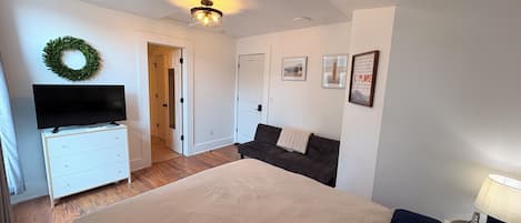 A bedroom with a bed, a television on a dresser, a dark sofa, a wall-mounted light fixture, and a door leading to another room.