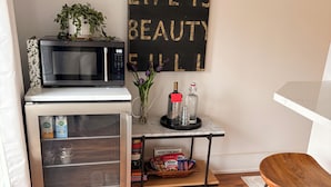 Cocina compacta con microondas, refrigerador y una mesa pequeña con aperitivos y una botella. Un cartel motivacional está en la pared.