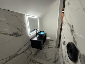 A bathroom with a marble floor, a black sink, a white toilet, and a mirror with a light.