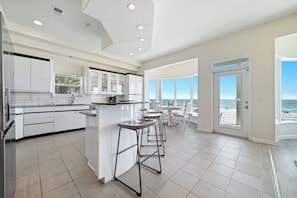 A modern kitchen with white cabinets, a central island, and bar stools. There is a large window with a view of the ocean.