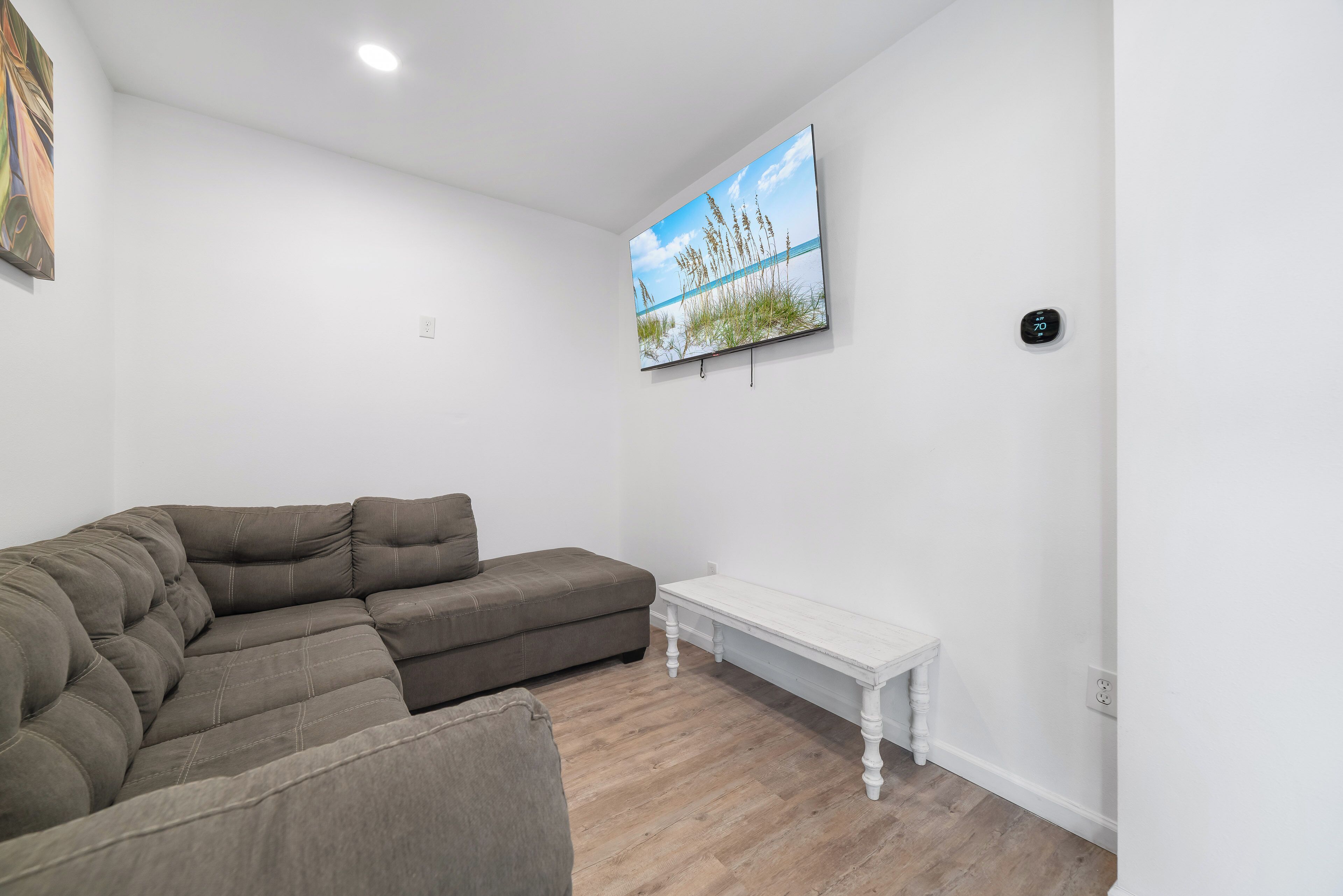 Un salon avec un canapé d’angle, une table basse blanche et un téléviseur fixé au mur qui diffuse une scène de plage.
