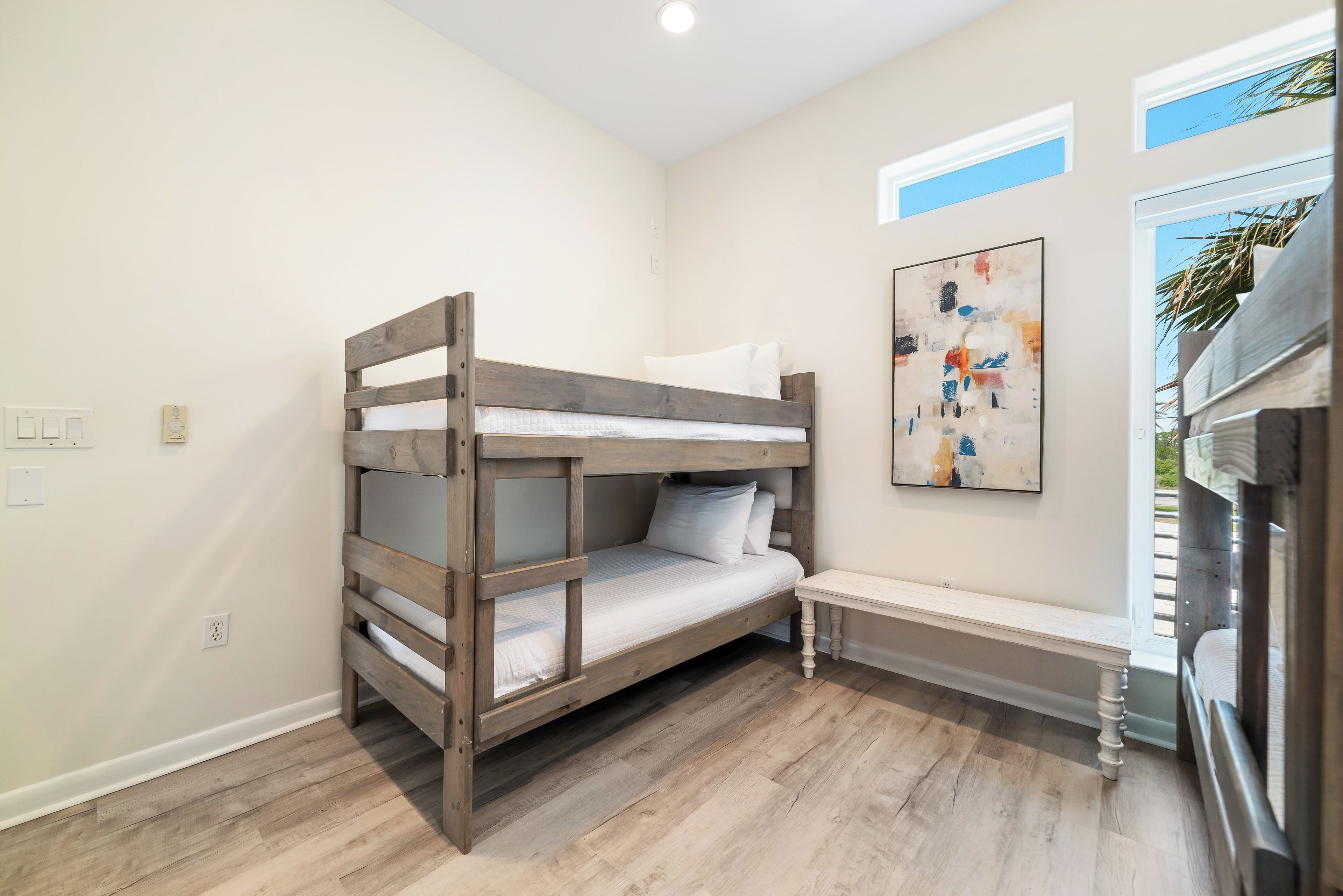 Una cama litera en una habitación con un banco, un cuadro colgado en la pared y una ventana con persianas.