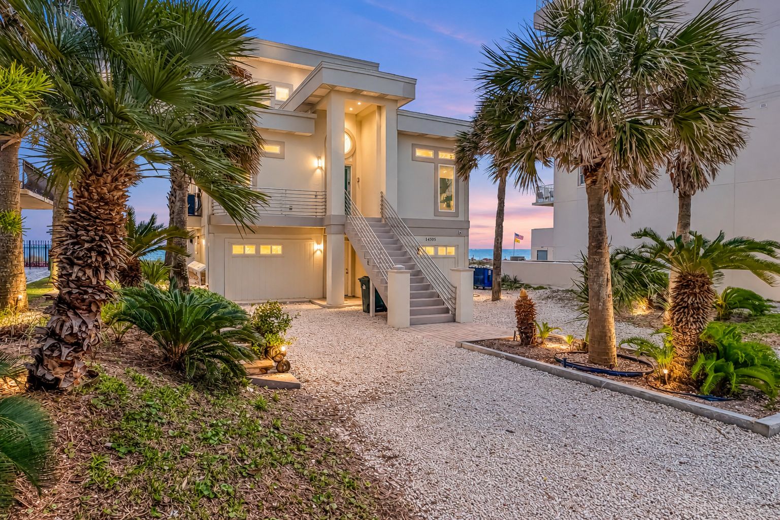 A modern two-story building with a central staircase, surrounded by palm trees and a gravel driveway.