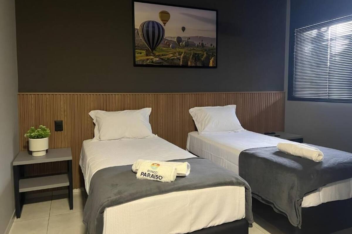 A hotel room with two beds, a wooden headboard, a framed picture on the wall, a small table with a plant, and a window with blinds.