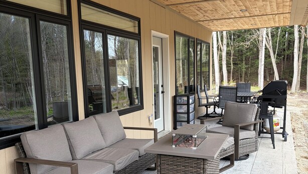 Ein überdachter Patio-Bereich mit Korbmöbeln, einem Grill und großen Fenstern mit Blick auf ein bewaldetes Gebiet.