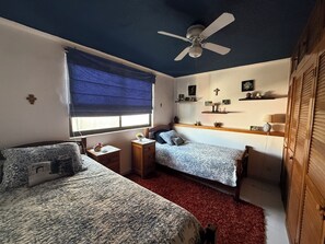 Une chambre avec deux lits, un ventilateur de plafond, une fenêtre avec des stores, une moquette rouge et une armoire en bois.