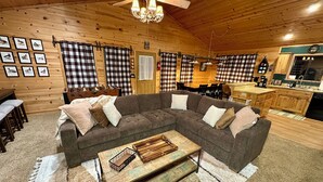 A cozy cabin interior with a sectional sofa, wooden flooring, a dining area, and a kitchen. - Greer Mountain Cabin: Scenic Deck, Rustic Charm & Stress-Free Parking (Greer)