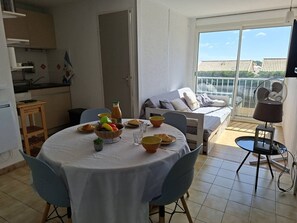 A dining area with a table set for two, a kitchen in the background, and a balcony with a view. - Two-room apartment with swimming pool (SAINT PIERRE LA  MER)