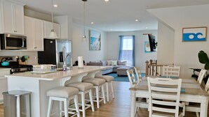 A modern kitchen with white cabinetry, a central island with bar stools, a dining area with white chairs, and a living room with a sofa and a TV. - Bayside Break | Rooftop Deck + Resort Pools & Gym (Berlin)
