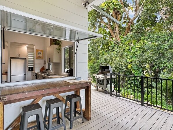 An outdoor deck with a wooden table and bar stools, a barbecue grill, and a view of a lush garden.