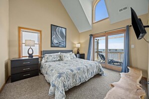 A bedroom with a bed, a nightstand, a lamp, a window, a painting, a balcony with a view, and a ceiling fan.