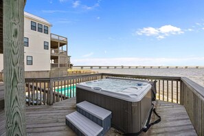 A wooden deck with a hot tub, a pool, and a view of the beach and pier.