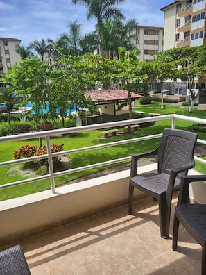 A balcony with a view of a swimming pool, palm trees, and a building with a tiled roof. - Beautiful Costa Linda beach condo, 2 BR/ 2 BA, Pool view (Jacó)