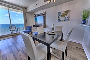 A modern dining area with a large table, chairs, a TV, and a view of the ocean. - Sunset Serenity 1302-Gulf Views-Sleeps 6 (Panama City)