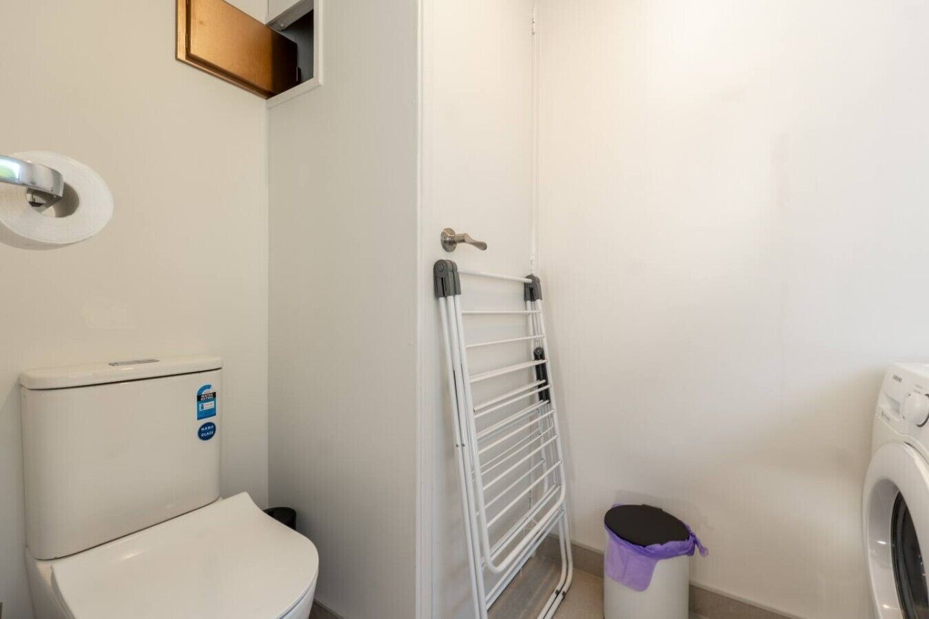 A bathroom with a toilet, a white wall, a metal drying rack, a trash bin, and a washing machine.