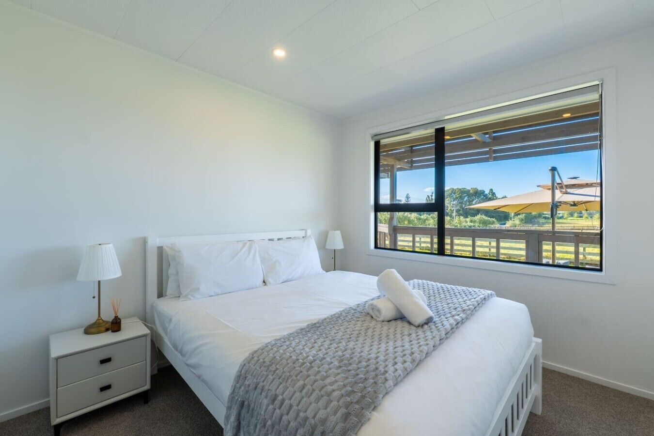 A bedroom with a bed, nightstand, lamp, and a window with a view of a deck and greenery.