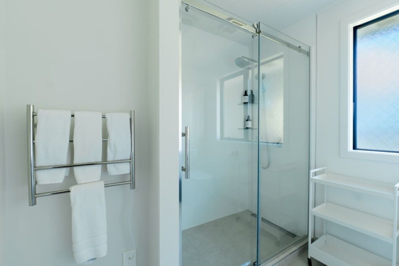 A modern bathroom with a glass-enclosed shower, a towel rack with multiple towels, and a window providing natural light.