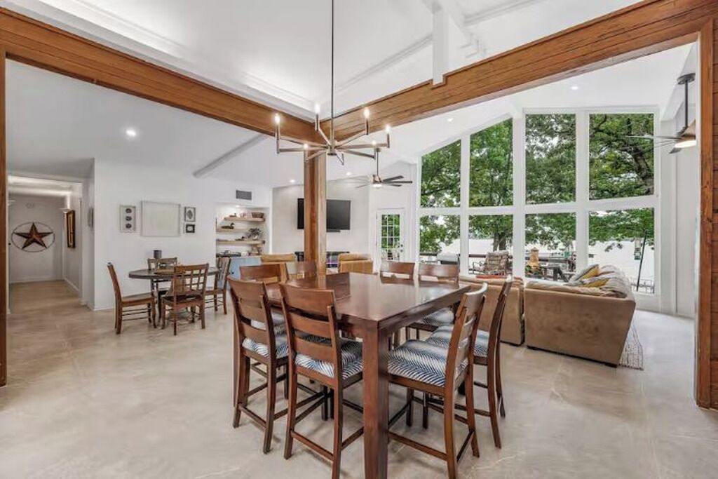 Un vaste espace dédié à la salle à manger, avec une grande table et des chaises en bois, une cuisine à l’arrière-plan et de grandes fenêtres offrant une vue sur les arbres.
