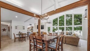 Un vaste espace dédié à la salle à manger, avec une grande table et des chaises en bois, une cuisine à l’arrière-plan et de grandes fenêtres offrant une vue sur les arbres.