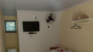 Ein Zimmer mit einem Bett, einem Fernseher, einem Ventilator, einem kleinen Tisch mit Stuhl und einem Fenster.
