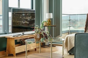 Una habitación moderna con un ventanal grande, un escritorio con computadora, una mesa de vidrio, un sofá y una planta.