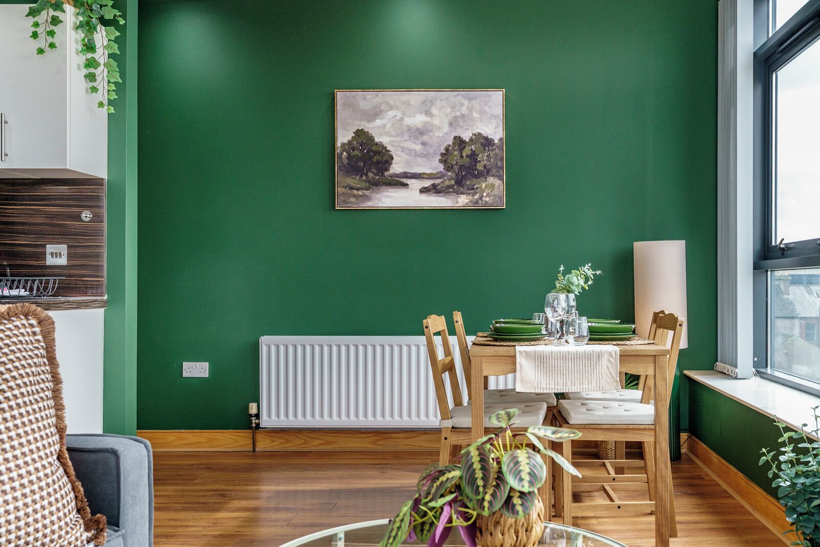 A dining area with a green wall, a painting, a radiator, wooden chairs, a table, and a window.