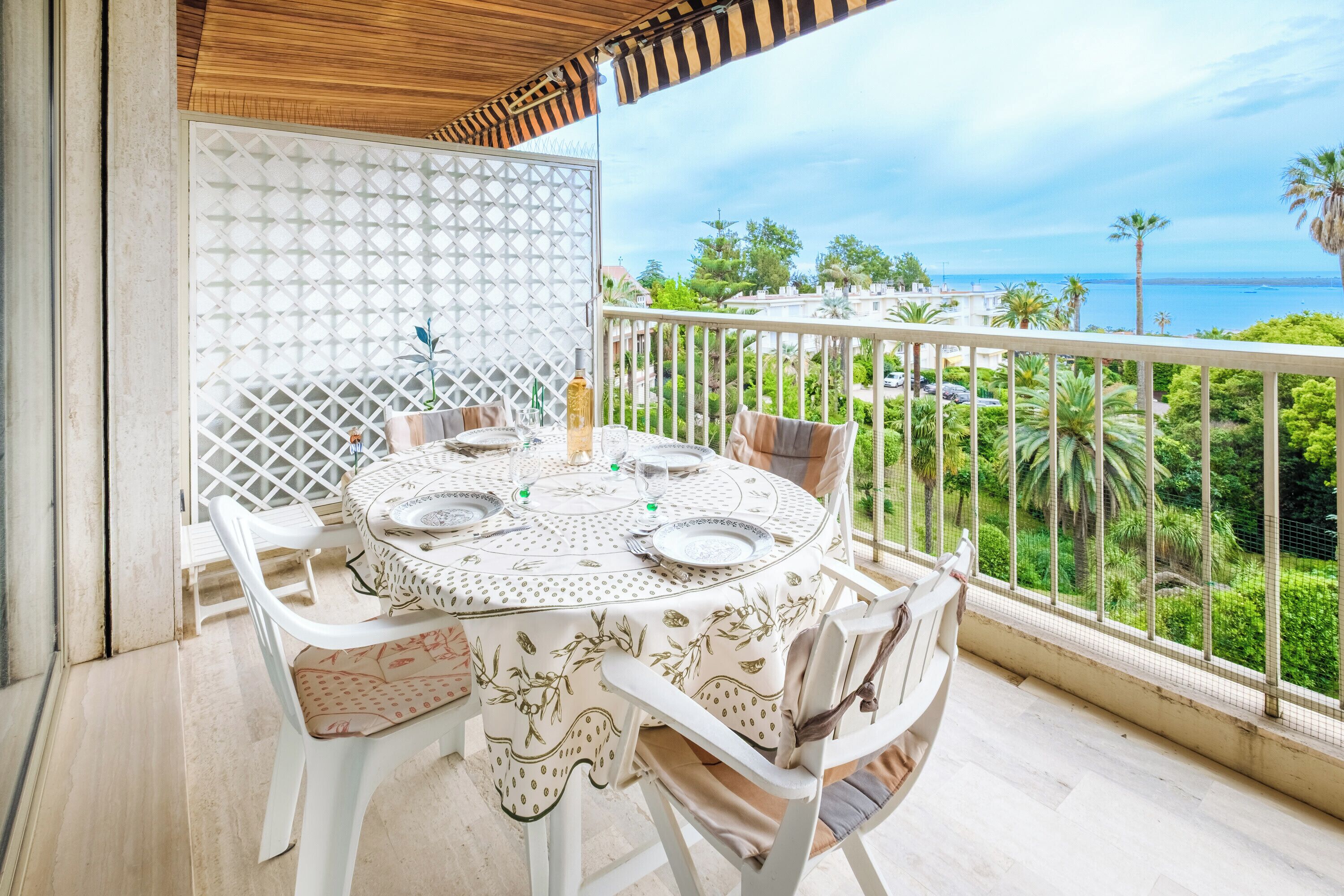 Une table ronde entourée de chaises blanches et recouverte d’une nappe à motifs, installée sur un balcon offrant une vue sur la côte, avec des palmiers et un ciel dégagé.