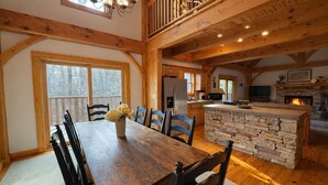 A spacious dining area with a wooden table and chairs, a stone island in the kitchen, and a large window with a view of trees. - Peaceful Resort Cabin in Sapphire Valley close to Cashiers & resort access (Sapphire)