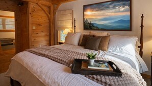 A cozy bedroom with a wooden ceiling, a large bed with white linens, a painting of a sunset, and a bedside table with a lamp. - Peaceful Resort Cabin in Sapphire Valley close to Cashiers & resort access (Sapphire)