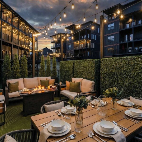 An outdoor dining area with string lights, a fire pit, and a green hedge wall.