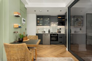 A modern kitchen with grey cabinets, a dishwasher, and an oven. There is a dining area with a table and chairs. - The Valour | Designer Apartment with Balcony (Cape Town)