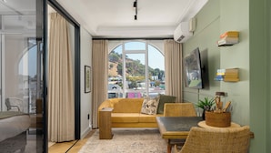 A room with a sofa, a dining table, a TV, and a view of a hillside. - The Valour | Designer Apartment with Balcony (Cape Town)