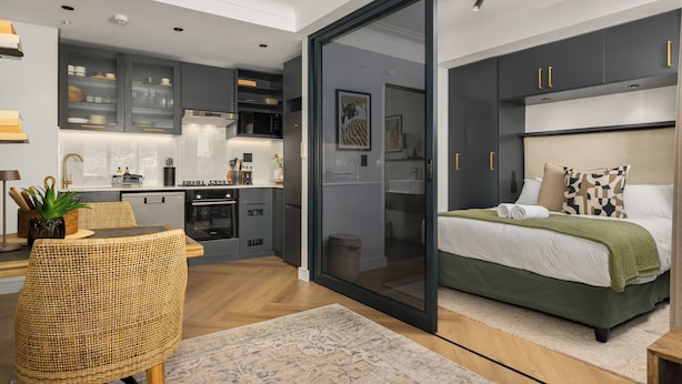 A modern bedroom with a bed, a dining area with a table and chairs, a kitchen with cabinets and appliances, and a sliding glass door leading to another room. - The Valour | Designer Apartment with Balcony (Cape Town)