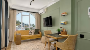 A living room with a sofa, a dining table with chairs, a flat-screen TV, and a view through a large window. - The Valour | Designer Apartment with Balcony (Cape Town)
