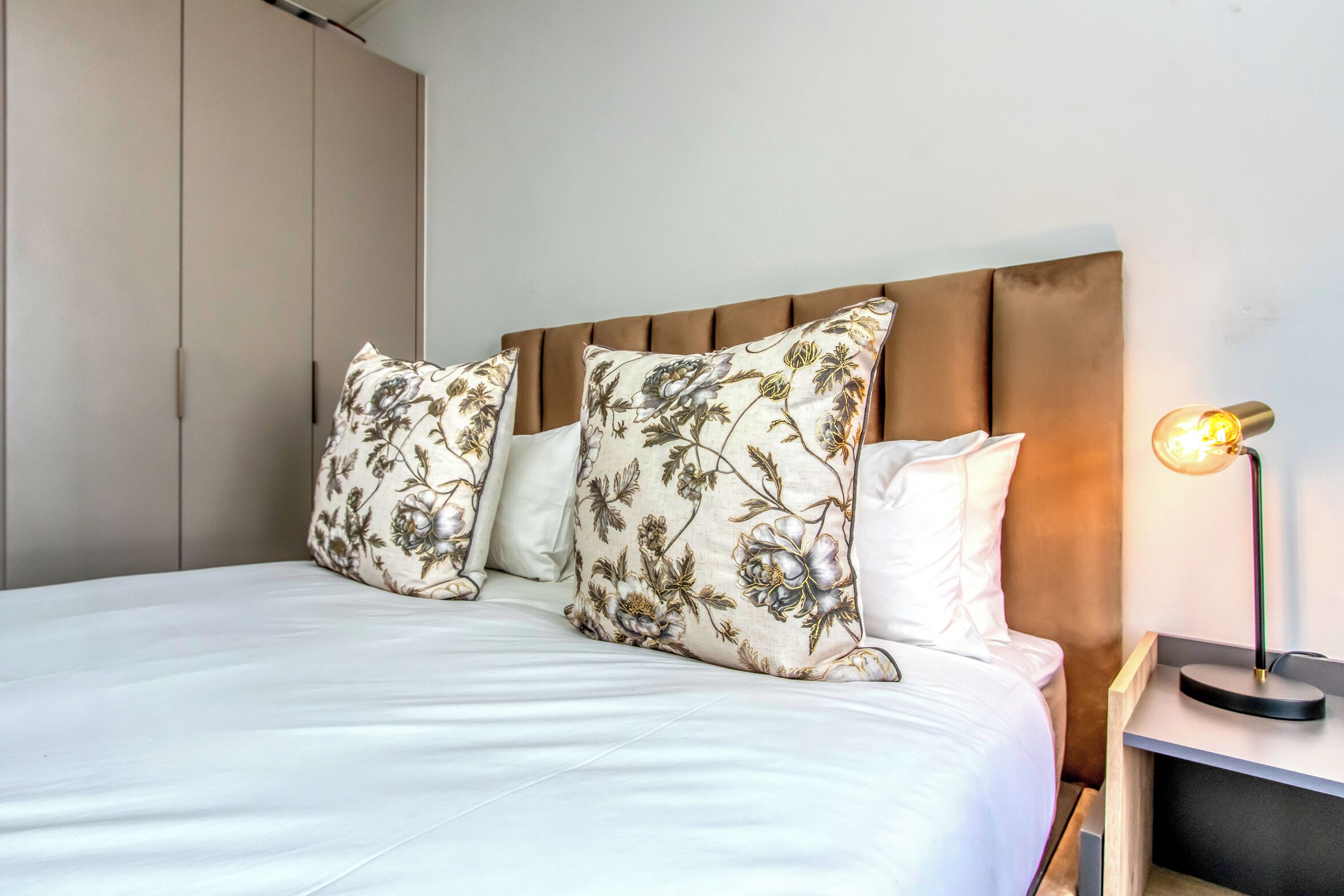 A neatly made bed with floral-patterned pillows, a wooden headboard, and a bedside lamp.