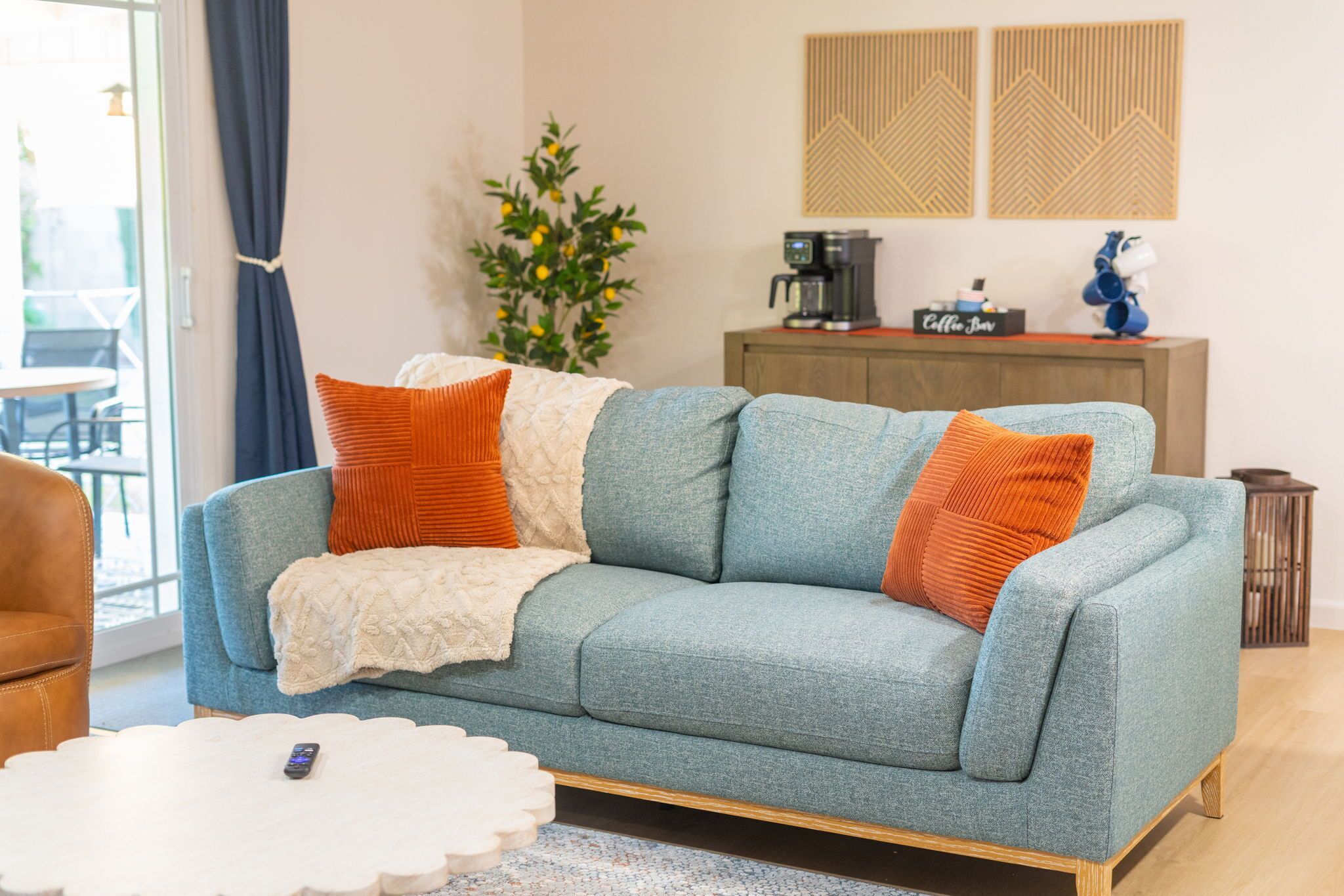 A living room with a blue sofa, orange cushions, a white coffee table, and a coffee machine on a wooden cabinet.