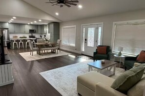 Eine moderne Küche mit grauen Schränken, eine Essecke mit Tisch und Stühlen, ein Wohnzimmer mit Sofa und Couchtisch sowie ein Deckenventilator.