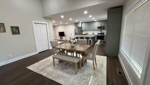 Een moderne keuken met inbouwapparatuur, een eethoek met een houten tafel en bank, en een groot raam met zonwering.