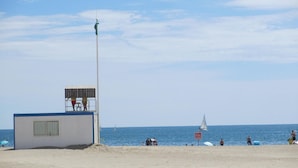 Una torre de socorrista con una bandera en la parte superior, una playa con gente y un velero a lo lejos.
