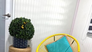 A yellow wicker chair with a teal cushion, a potted plant with yellow flowers, and woven mats on a wooden floor. - Cozy 2-Bedroom Retreat in Cabo Rojo Near the Beach (Cabo Rojo)