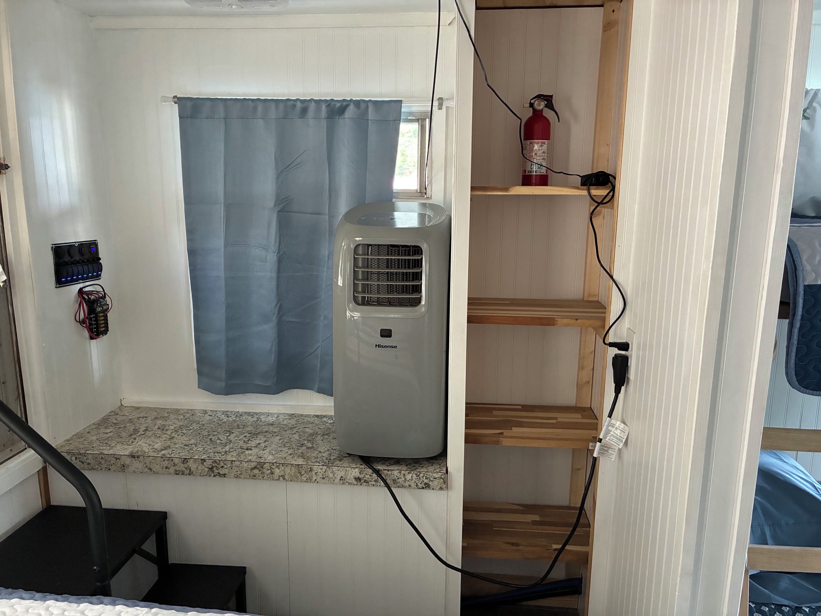 A compact room with a Hisense air conditioner, a fire extinguisher on a shelf, and a window with a blue curtain.